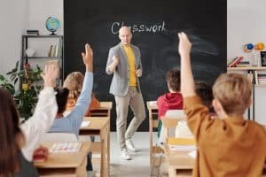 Teacher in a classroom in front of a blackboard, students raising their hands